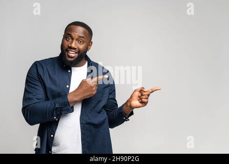 Positif et souriant Afro-américain 30s gars de pointe à côté de l'espace vide de copie, le type multiethnique présentant la nouveauté, isolé sur fond gris, offre de publicité concept, meilleure affaire Banque D'Images