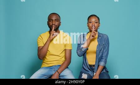 Homme et femme faisant le geste de silence avec l'index sur la bouche, demandant le secret et le calme.Couple montrant le signe de la vie privée pour garder le secret et demande de fermer, assis sur fond bleu Banque D'Images