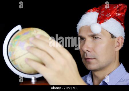 L'homme dans le chapeau de Santa tourne le globe avec sa main, portrait masculin sur fond noir.Concept de choisir le voyage de voyage pendant Noël et le nouvel an Banque D'Images