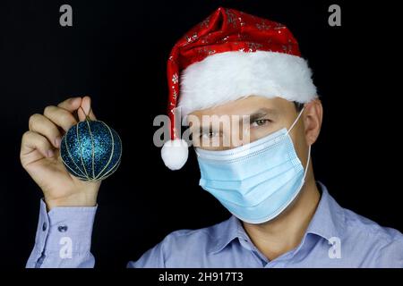 Fête de Noël, portrait d'homme en masque et chapeau de père Noël avec une balle de jouet bleue à la main.Concept de décoration de maison ou de bureau pour la nouvelle année Banque D'Images