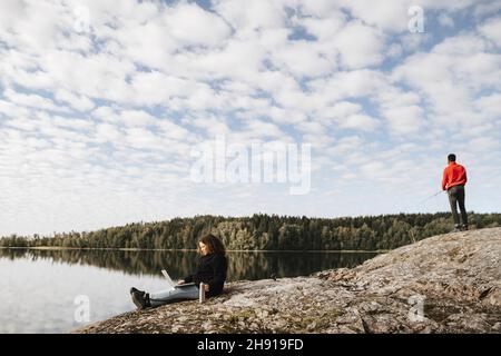 Femme travaillant à distance sur ordinateur portable pendant que l'ami pêche sur le bord du lac Banque D'Images