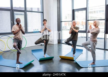 Les personnes âgées interraciales debout dans un arbre posent sur des tapis de yoga dans un centre sportif Banque D'Images