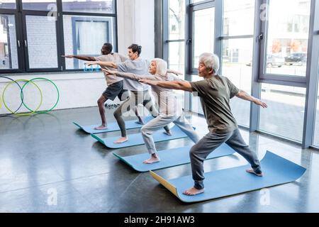 Un homme asiatique senior en guerrier pose pratiquer le yoga près d'amis multiethniques dans le centre sportif Banque D'Images