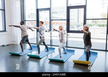 Les personnes âgées multiculturelles debout dans le guerrier posent sur des tapis dans le centre sportif Banque D'Images