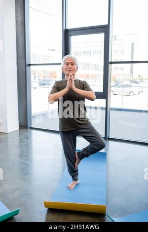 Un homme asiatique âgé debout dans un arbre pose sur un tapis de yoga dans un centre sportif Banque D'Images