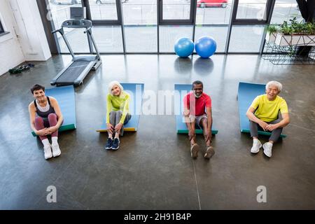 Vue en grand angle des personnes âgées multiethniques assises sur des tapis de fitness dans la salle de gym Banque D'Images