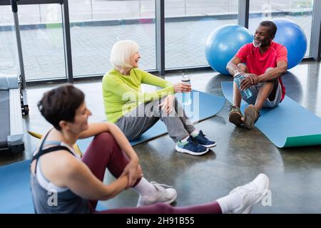 Des personnes âgées et interraciales gaies sont assises sur des tapis de fitness dans le centre sportif Banque D'Images