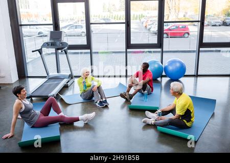 Joyeux senior interracial tenant des bouteilles de sport tout en étant assis sur des tapis de fitness dans la salle de gym Banque D'Images