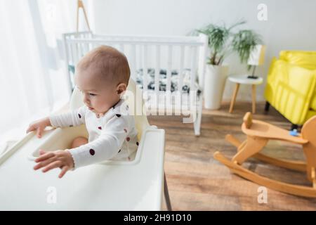 enfant assis dans une chaise de bébé près d'un berceau flou et d'un cheval à bascule Banque D'Images