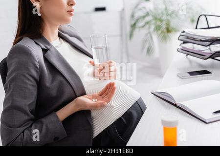 vue partielle de la femme enceinte tenant des pilules et un verre d'eau dans le bureau Banque D'Images