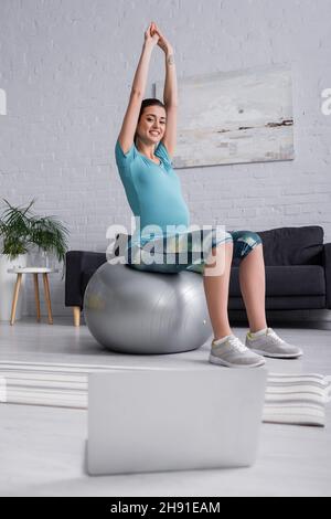 femme enceinte positive aux mains levées s'exerçant sur le ballon de fitness et regardant un ordinateur portable dans le salon Banque D'Images