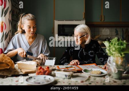 Une femme de famille coupe du pain à la table à manger tout en étant assise par une femme âgée dans la cuisine Banque D'Images