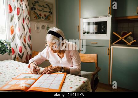 Femme senior résolvant le sudoku à la table de salle à manger dans la cuisine Banque D'Images