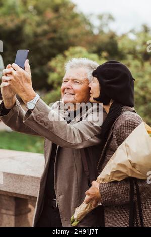 Une femme embrassant un homme âgé qui prend son selfie à travers un smartphone pendant le week-end Banque D'Images