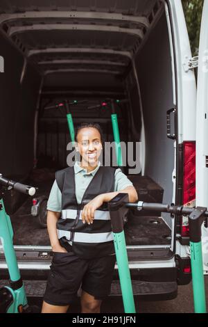 Femme souriante de livraison avec la main dans la poche penchée sur le scooter électrique pousser contre la camionnette Banque D'Images