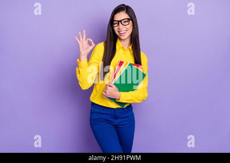 Photo de attrayante gaie jeune femme faire signe ok d'accord sourire tenir des livres isolés sur fond violet de couleur Banque D'Images