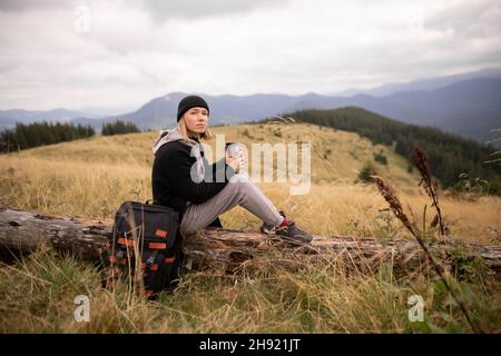 Jeune femme avec une coupe thermos reposant sur un arbre tombé dans les montagnes.Liberté, nourriture, voyage et vacances concept, activités de plein air. Banque D'Images