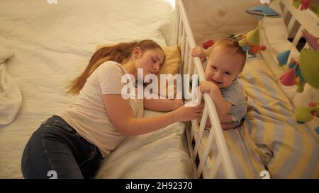 Pleurer et crier bébé garçon assis dans le lit tandis que la mère fatiguée dormant à côté de lui dans le lit Banque D'Images