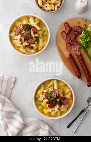 Soupe de pommes de terre et de pois jaunes avec croûtons et viandes fumées dans des bols, planche à découper avec oignons verts hachés et saucisses fumées, vue de dessus Banque D'Images