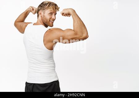 Vue arrière d'un beau sportif montrant ses muscles sur les bras, fléchissant les biceps et regardant un arrière-plan fort et blanc Banque D'Images