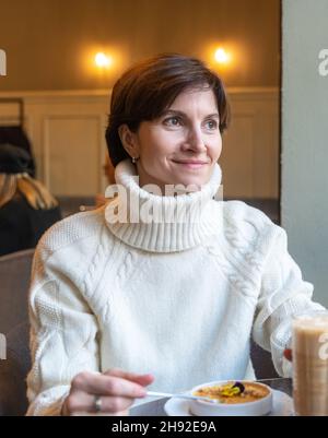 Femme souriante qui mange de la crème brûlée au café.Regarder la fenêtre.Vêtements d'automne Banque D'Images