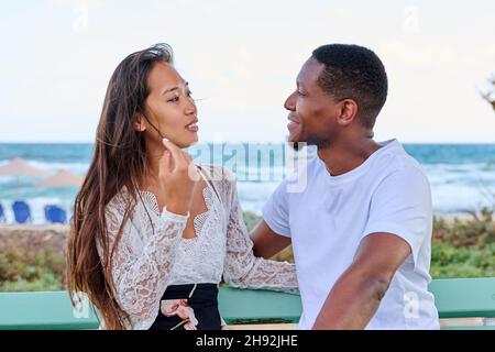 Jeune couple multiculturel souriant parlant assis sur un banc, fond de mer Banque D'Images