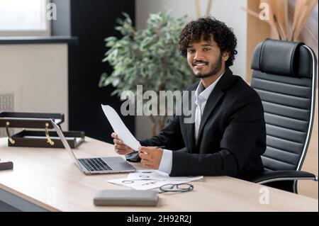 Portrait d'un homme d'affaires indien aux cheveux mauriques positif, vêtu d'une tenue d'affaires, assis à son bureau, regardant la caméra avec un sourire amical, détient des graphiques d'affaires Banque D'Images