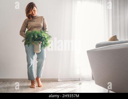 Une jeune femme vêtue chaud confortable chandail tenant le vase en argile rempli de branches vertes de sapin fraîchement coupé conifères près d'un mur blanc dans la maison liv Banque D'Images