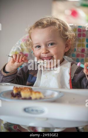 Une petite fille de 2 ans mange un peu de gâteau et de crème glacée comme collation de l'après-midi sur une chaise haute. Banque D'Images