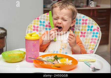 Une jeune fille de 2 ans grimace et pleure alors qu'elle mange un repas sur une chaise haute. Banque D'Images