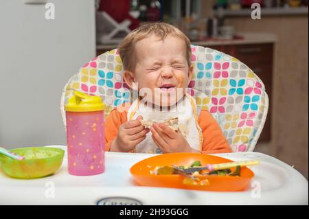 Une jeune fille de 2 ans grimace alors qu'elle mange un repas sur une chaise haute. Banque D'Images