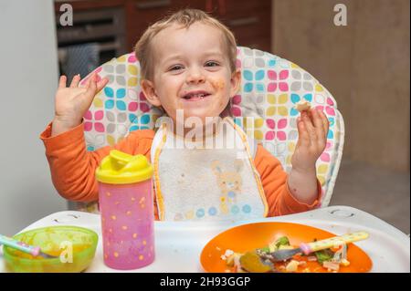 Une jeune fille de 2 ans sourit alors qu'elle mange un repas sur une chaise haute. Banque D'Images