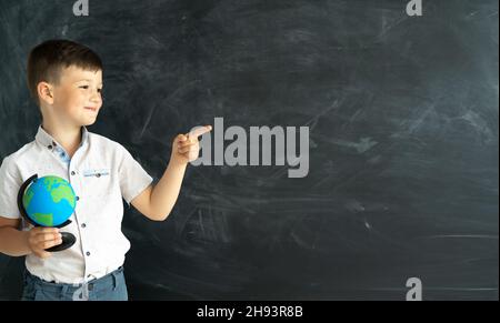 un écolier souriant à l'école avec un globe se tient devant un tableau de craie et pointe son doigt vers un endroit pour le texte.Concept d'éducation et d'étude Banque D'Images