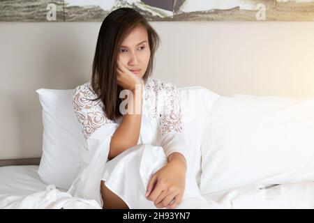 Après une nuit de sommeil triste, la femme s'est assise sur le lit en pensant.Femme asiatique en dentelle blanche de nuit est l'insomnie Banque D'Images