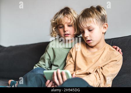 Un petit frère calme et un cheveu blond dans des tenues décontractées pour regarder des vidéos intéressantes sur un smartphone, tout en étant assis ensemble sur un canapé confortable à la maison Banque D'Images