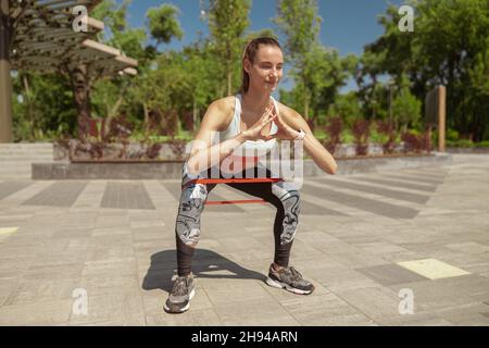 Femme souriante dans les vêtements de sport fait des squats avec un pli élastique dans le parc Banque D'Images