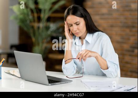 Exténuée femme caucasienne, directrice, secrétaire, fatiguée de travailler avec un ordinateur portable pendant qu'elle est assise sur son lieu de travail.Une femme d'affaires surtravaillée frotte le pont de son nez, tient des lunettes dans sa main, ont la migraine Banque D'Images