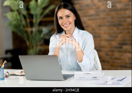 Portrait d'une jolie femme brune caucasienne.Une employée de bureau réussie, une directrice, une conseillère d'affaires, portant une chemise habillée, s'asseoir sur son lieu de travail avec un ordinateur portable, regarde la caméra, souriant amical Banque D'Images