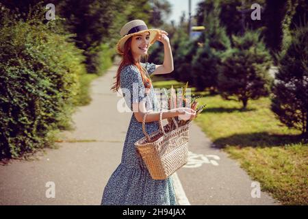 vue arrière de la jeune femme heureuse avec sac en paille élégant et chapeau en paille qui marche dans le parc à l'extérieur Banque D'Images