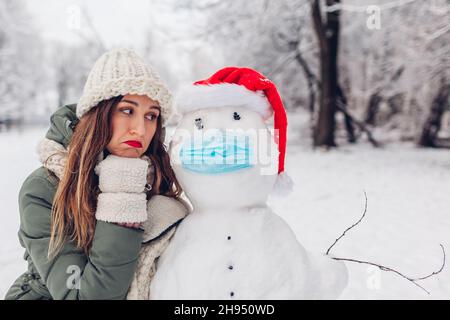 Une femme triste sympathise avec un bonhomme de neige malade portant un masque médical et un chapeau de père Noël dans un parc hivernal enneigé.Mesures de protection en cas de coronavirus covid pandem Banque D'Images