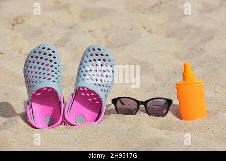 Gros plan sur des chaussures de sabots, de la crème solaire et des lunettes de soleil noires de protection sur une plage de sable au bord de la mer tropicale par temps chaud et ensoleillé. Concept de vacances d'été. Banque D'Images