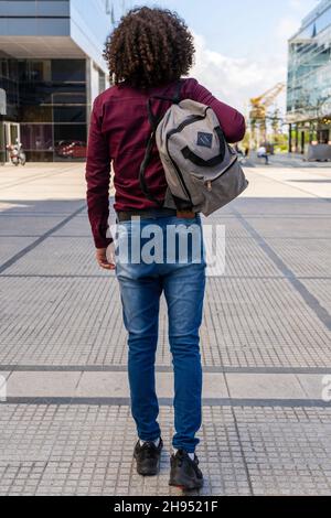 Un portrait complet de l'arrière d'un jeune homme avec un sac à dos qui marche en descendant la rue. Banque D'Images