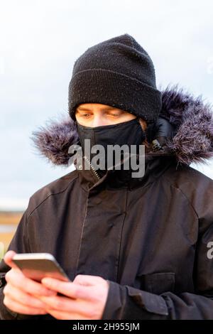 Un jeune homme portant un masque noir lavable tout en utilisant son téléphone portable pendant la pandémie du virus corona Covid-19 Banque D'Images