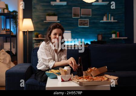 Portrait d'une femme souriante se reposant sur un canapé après un travail de bureau en appréciant un dîner de restauration rapide le soir.Menu de restauration rapide à emporter.Commande de nourriture indésirable sur la table basse. Banque D'Images