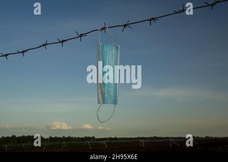 Image conceptuelle d'un masque chirurgical de protection sur un fil barbelé contre un fond de paysage. Banque D'Images