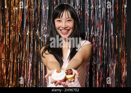 Une jeune femme asiatique montre des cupcakes à l'appareil photo sur un fond de fête. Banque D'Images