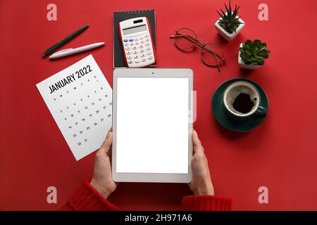 Les femmes ont des mains avec un ordinateur tablette, un calendrier papier pour janvier 2022, une calculatrice, des lunettes et une tasse de café sur fond de couleur Banque D'Images