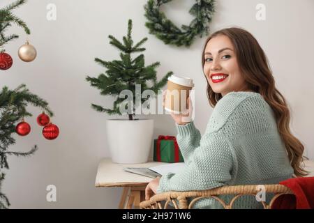 Belle femme souriante tenant une tasse en papier avec une boisson dans la chambre décorée pour Noël Banque D'Images