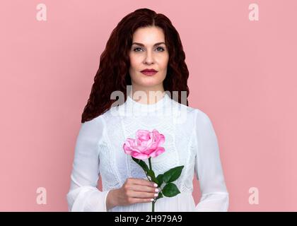 Femme avec une rose contre fond rose.Gros plan portrait d'une belle femme avec des cheveux bruns rouges ondulés tenant une rose rose délicatement. Banque D'Images