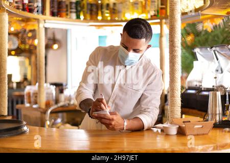 Jeune serveur latino-américain avec masque médical prenant des notes dans son carnet au bar d'un restaurant élégant.Espace pour le texte. Banque D'Images